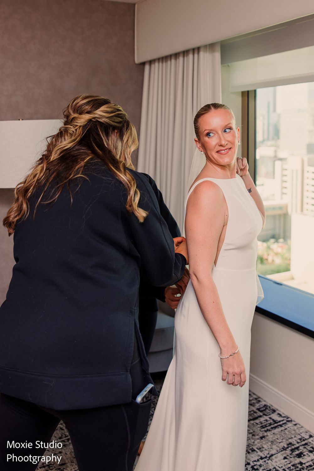 Bride in an elegant white sleeveless gown smiles over her shoulder as a woman in navy fastens the back in a bright hotel room.
