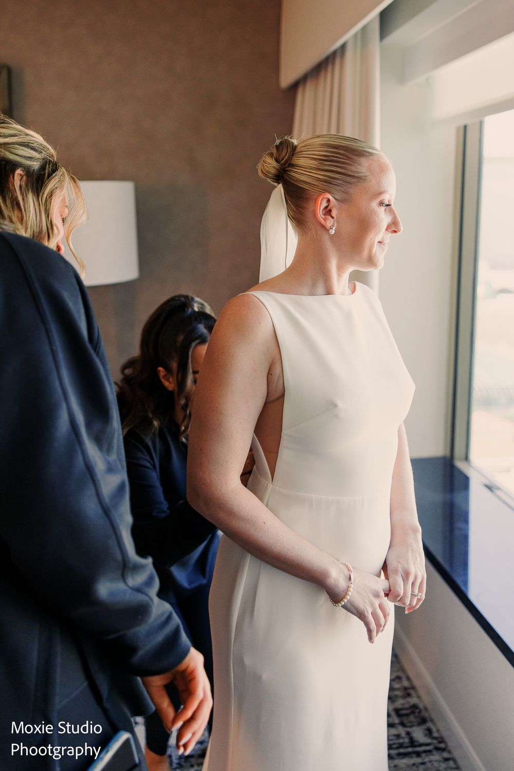 Bride in a sleek white sleeveless gown with a low bun and veil looks out a hotel window while bridesmaids help her get ready.