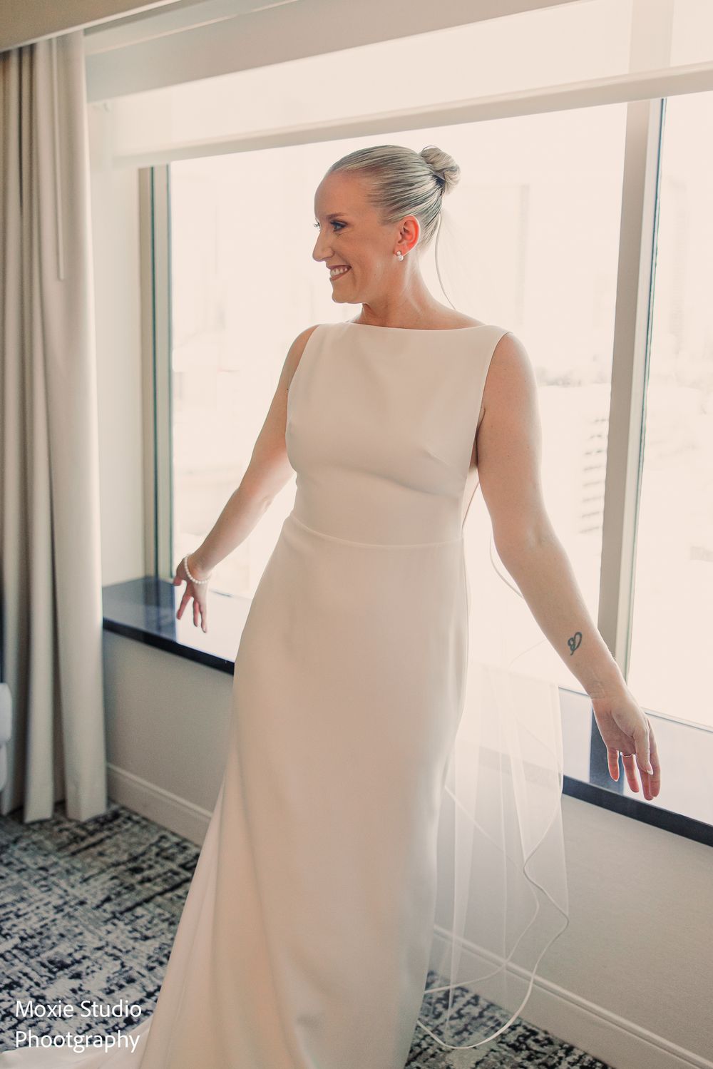 Smiling bride in an elegant sleeveless ivory wedding gown and sheer veil stands beside a large sunlit window in a hotel room.