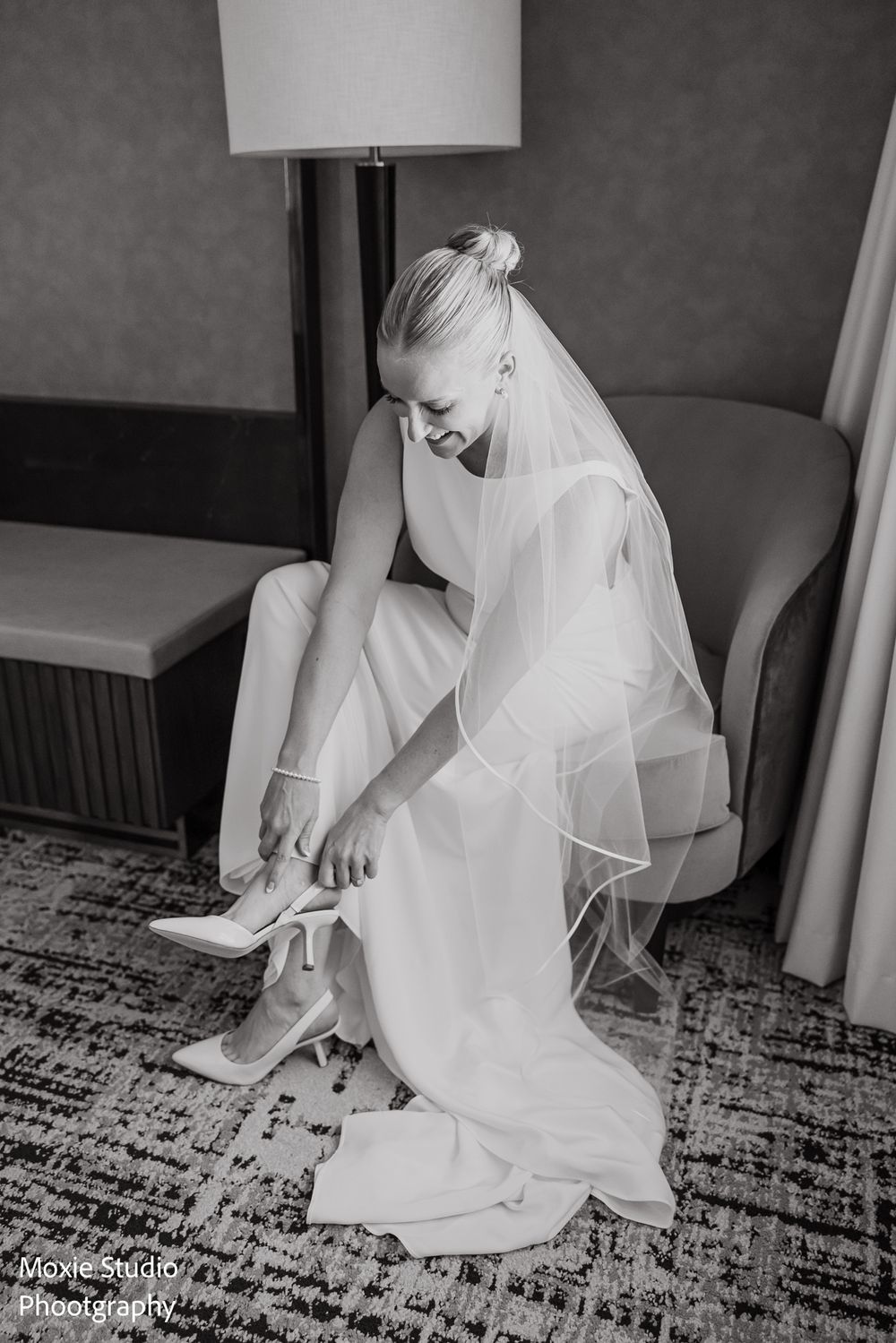 Black and white photo of a bride in a sleek gown and veil with hair in a bun, sitting in a chair while putting on her white heels.