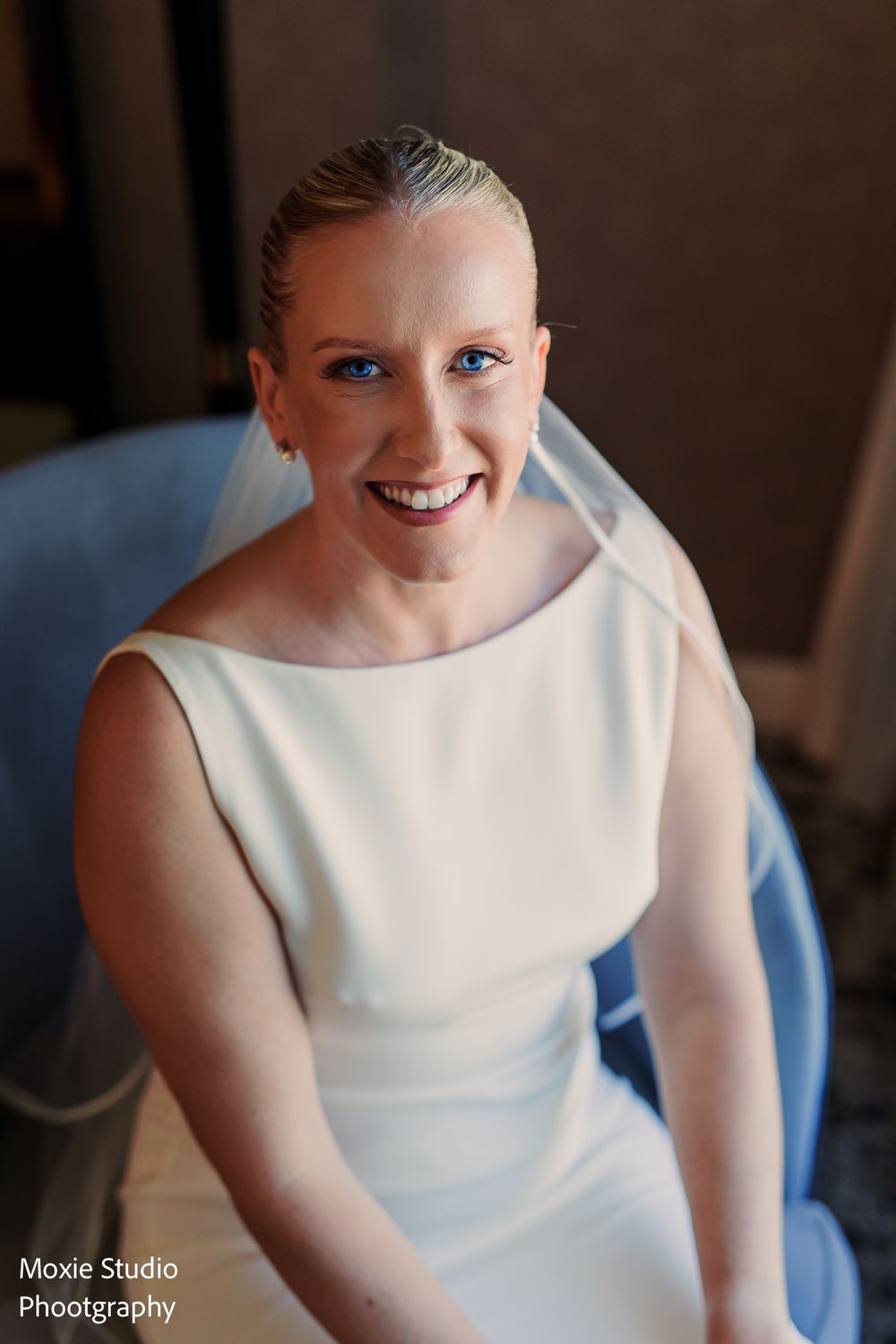Smiling bride with blue eyes sits in a blue chair wearing a sleek white sleeveless wedding gown and a sheer veil.