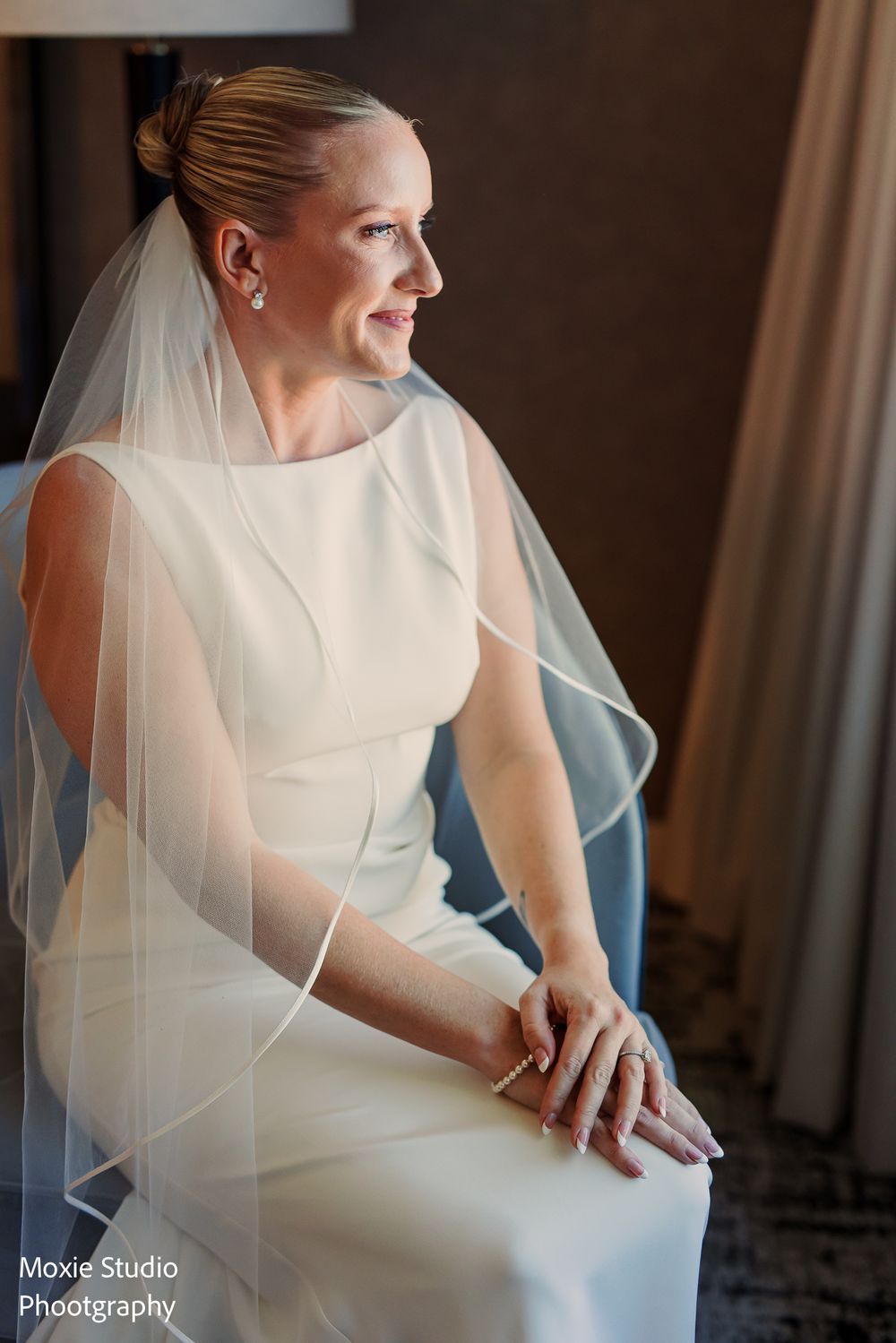 Bride in an elegant white sleeveless gown and sheer veil sits in a blue chair, gazing thoughtfully out a window.