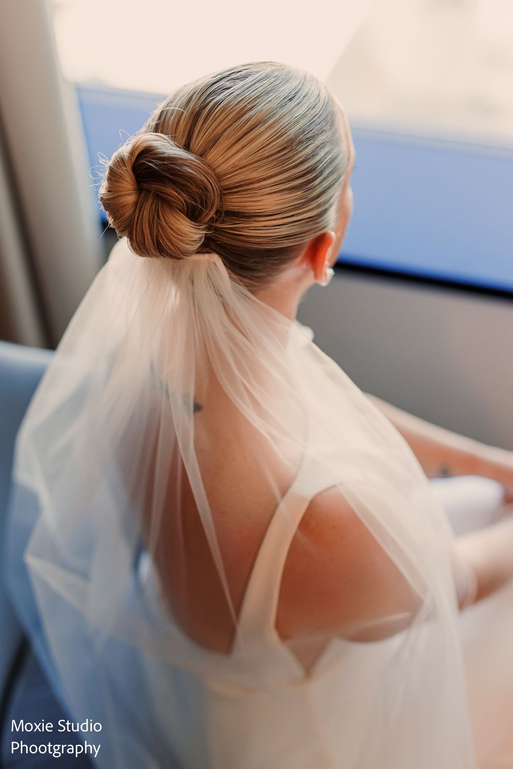 Bride in a white gown with a sleek low bun and sheer tulle veil gazes out a window in a romantic wedding portrait.