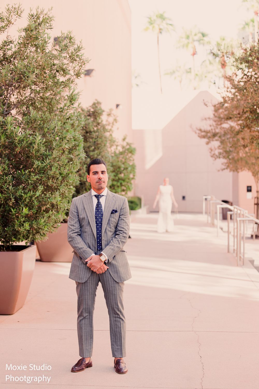 Groom in a gray pinstripe suit and navy tie stands outdoors while the bride in a white gown waits in the distance at a sunny wedding venue.