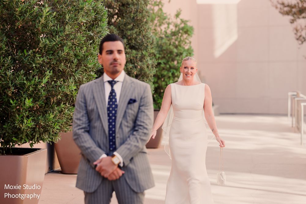Groom in a gray suit and navy polka dot tie stands with hands clasped as the bride in a white gown walks toward him in a first look.