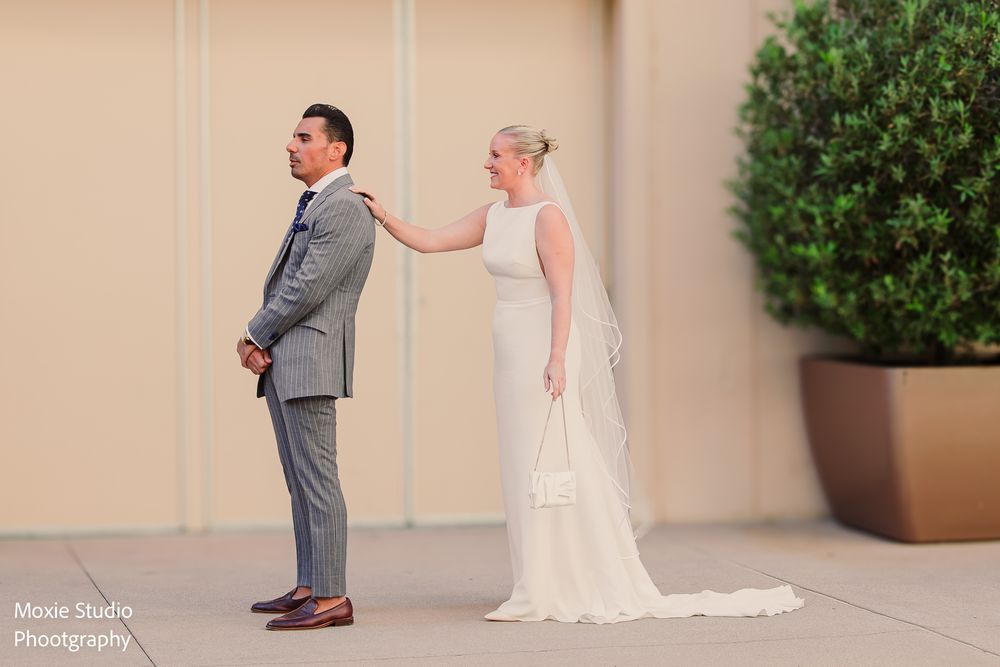 Bride in a white gown and veil taps the shoulder of a groom in a gray suit during a first look photo outdoors by a potted shrub.