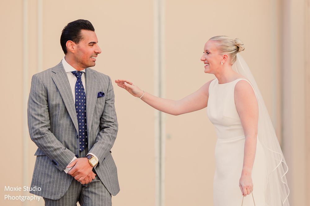 Bride in a white gown and veil reaches out toward the groom in a gray pinstripe suit during their joyful first look.