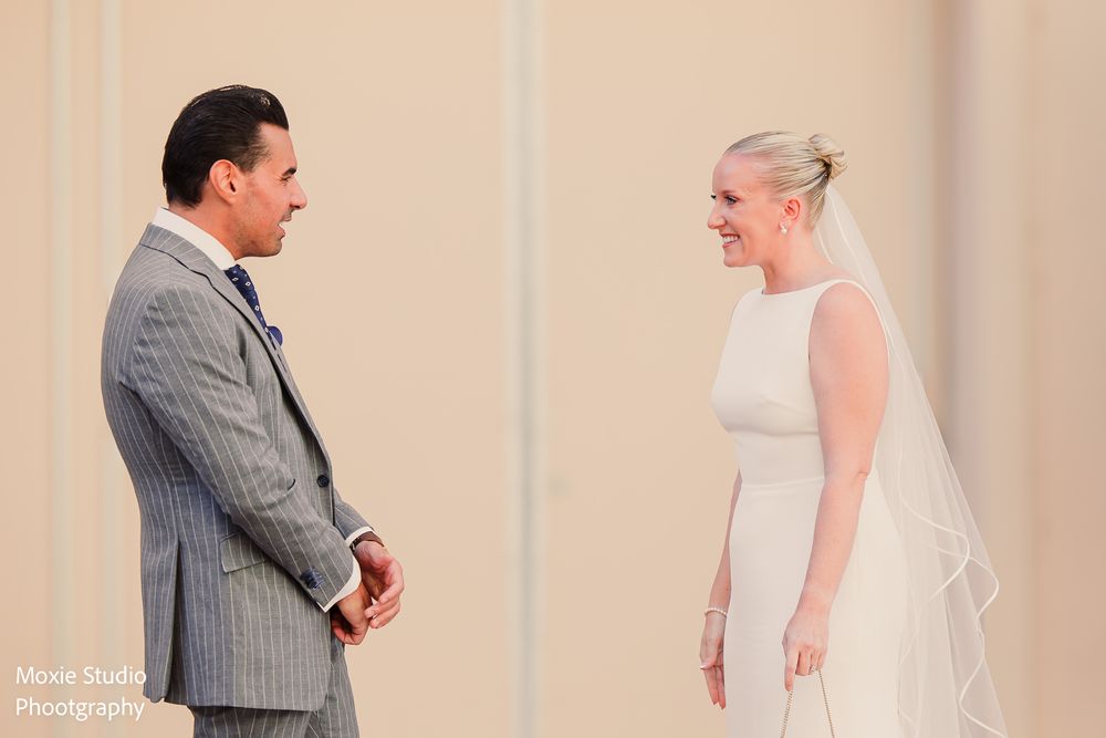 Groom in a gray pinstripe suit and bride in a white gown and veil smile at each other during an emotional wedding first look.
