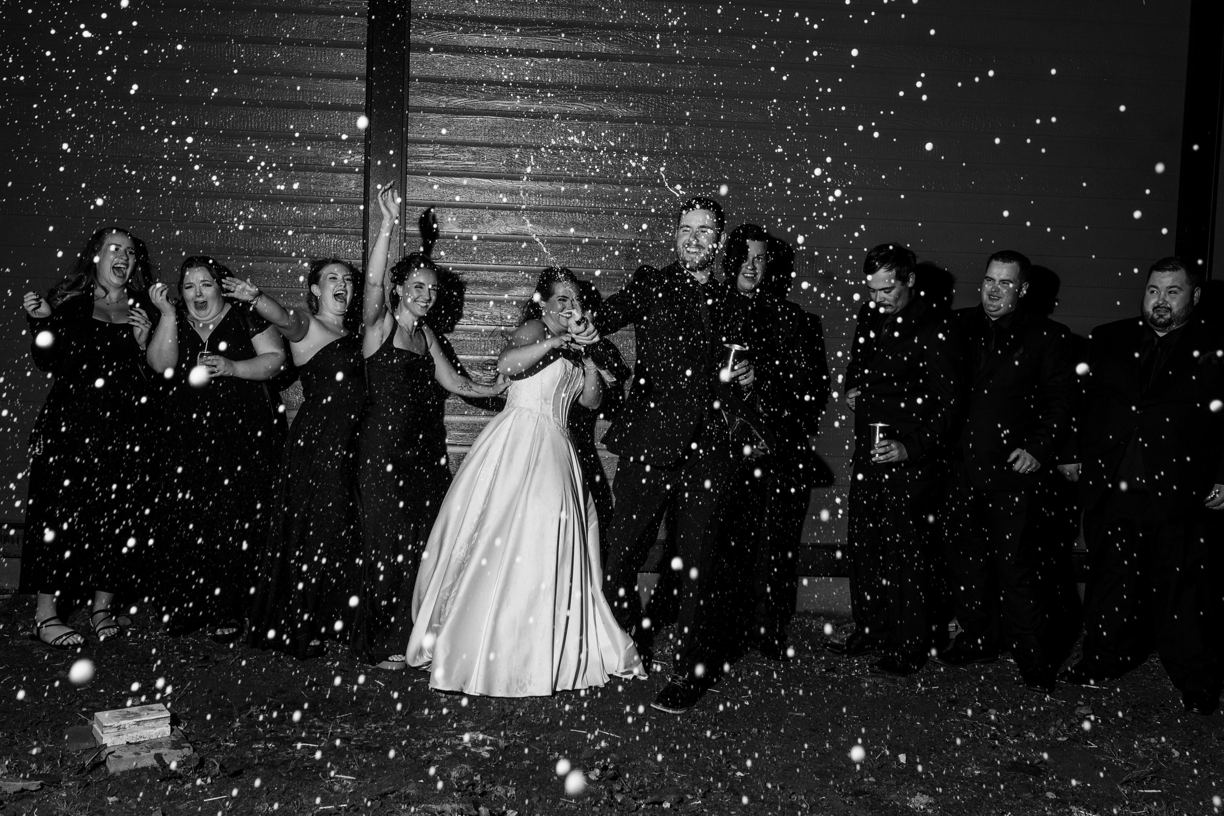 Black and white wedding photo of a bride in a white gown surrounded by cheering bridesmaids and groomsmen as confetti falls around them.