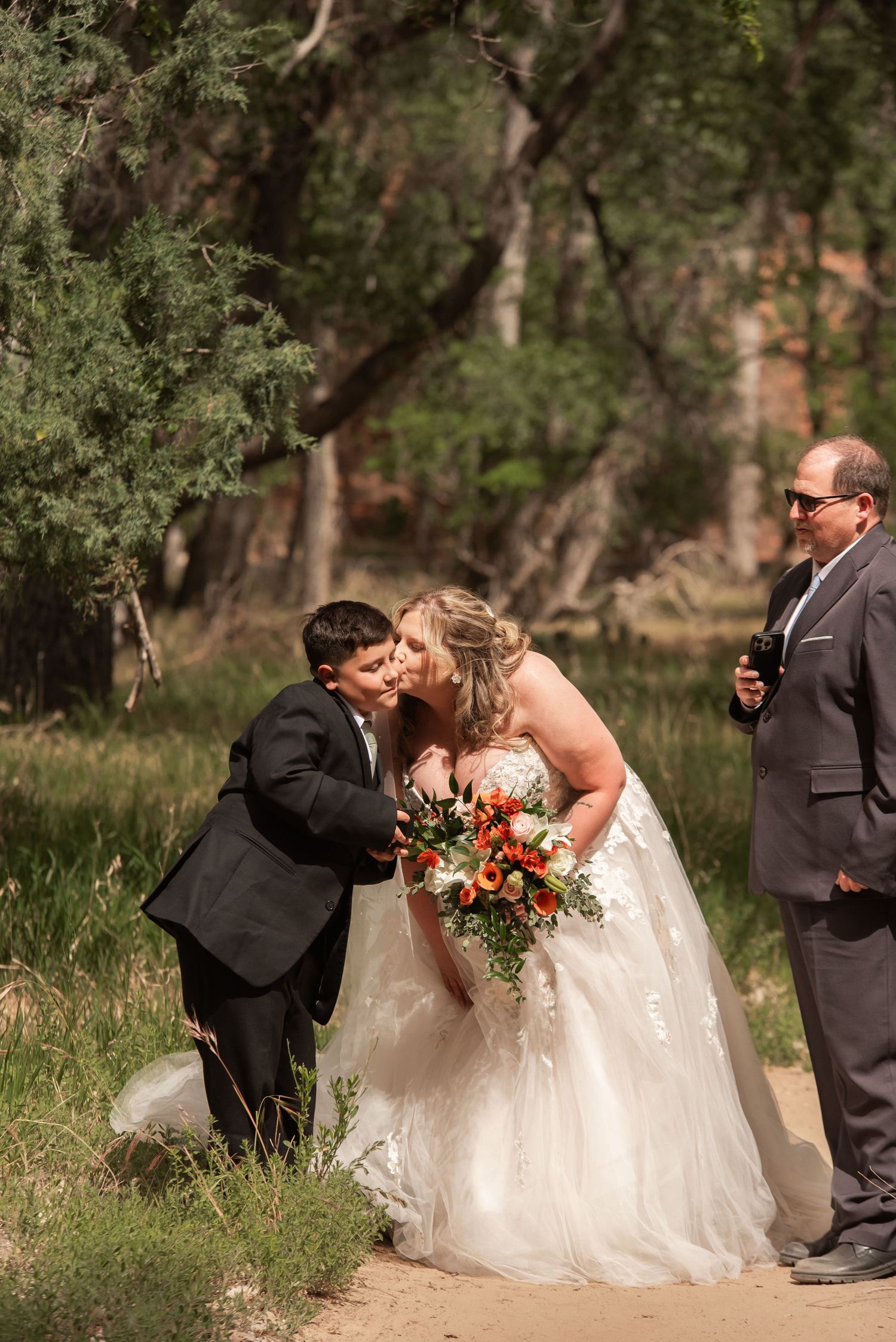 Bride in white gown kisses young boy in suit holding bouquet outdoors at woodland wedding ceremony.