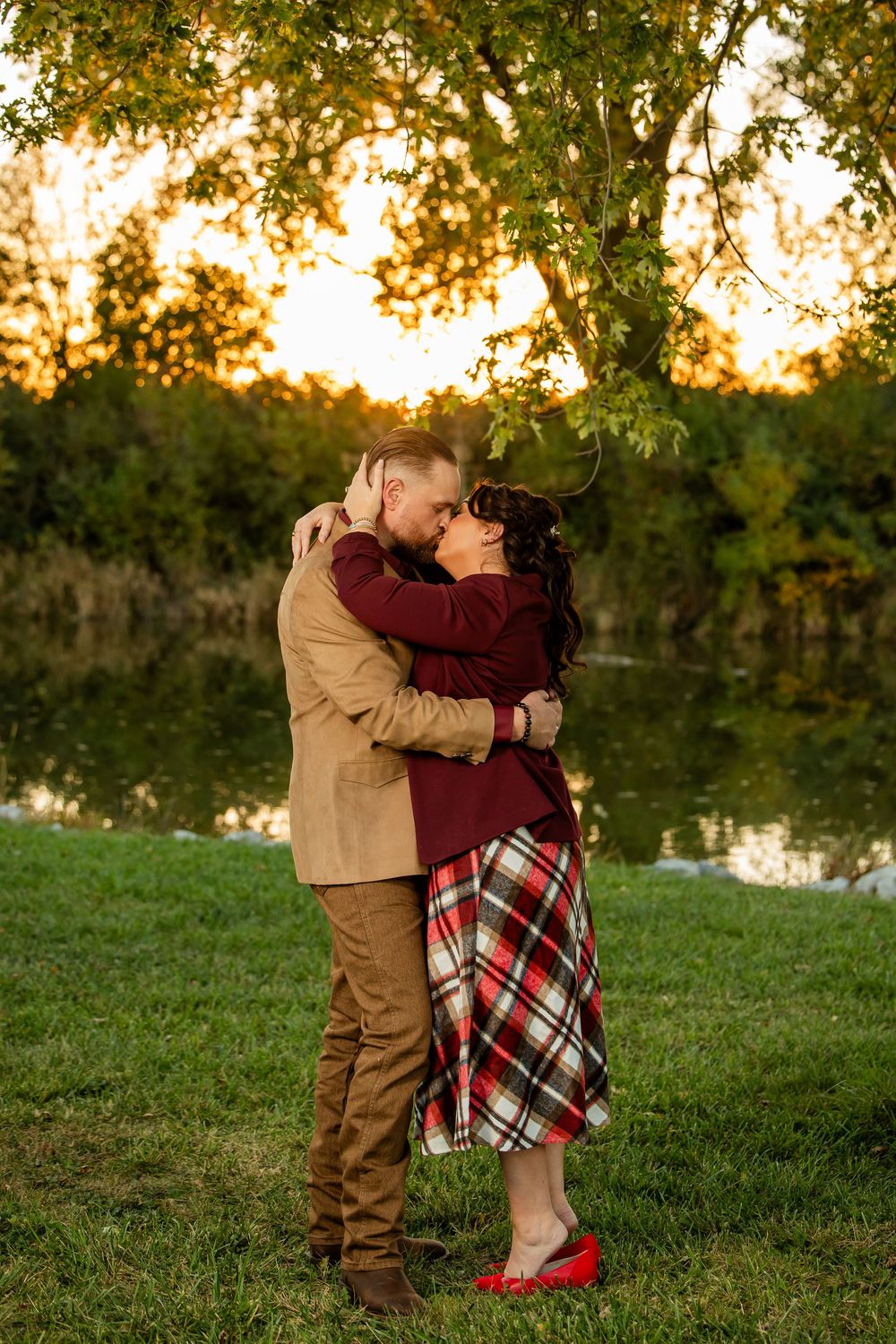 Couple embracing and kissing by a tranquil pond at golden sunset, surrounded by lush green trees in autumn.
