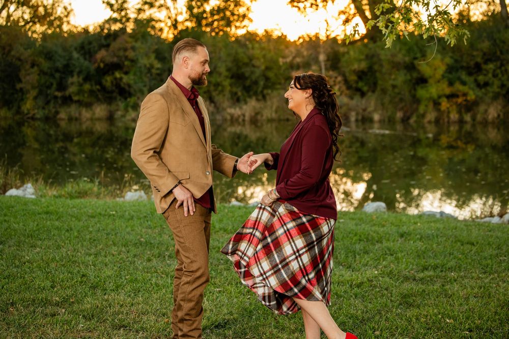 Couple holding hands and dancing outdoors near a reflective pond at golden hour, wearing autumn attire.
