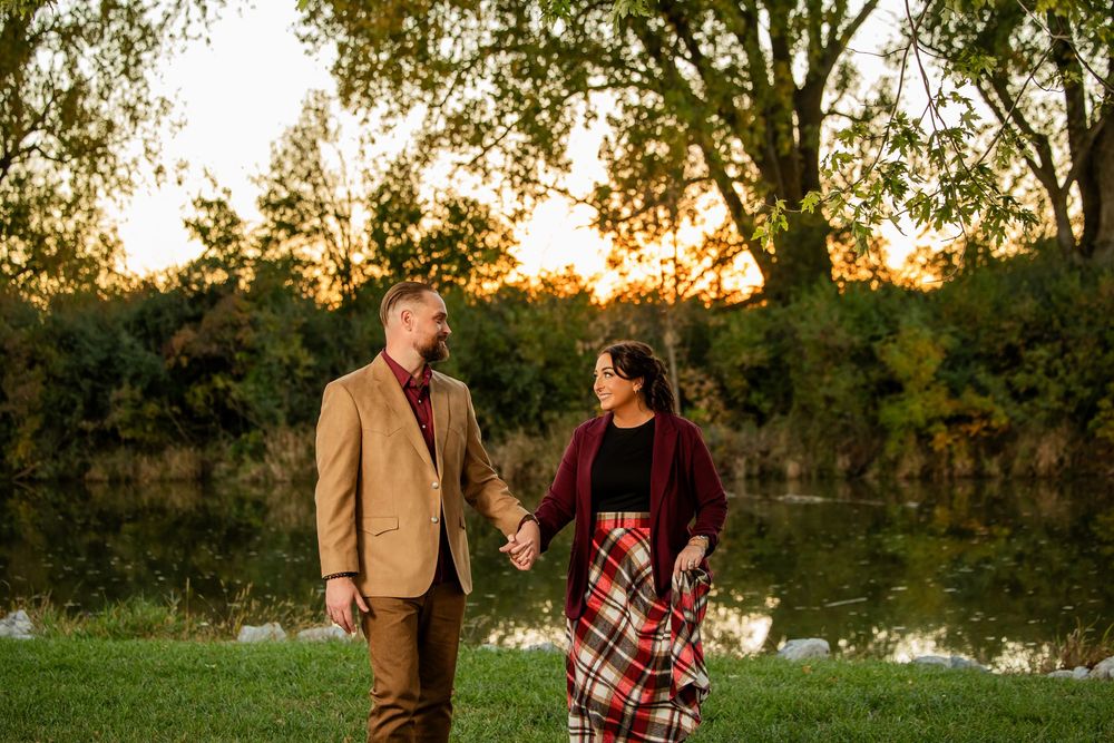 Couple holding hands at sunset by a pond, dressed in autumn colors, surrounded by golden trees.