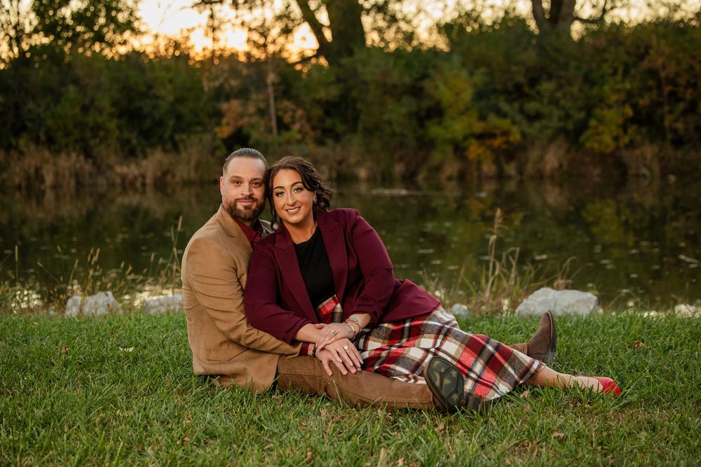 Couple sitting together on grass by a pond in autumn, smiling, wearing burgundy and tan outfits at sunset.