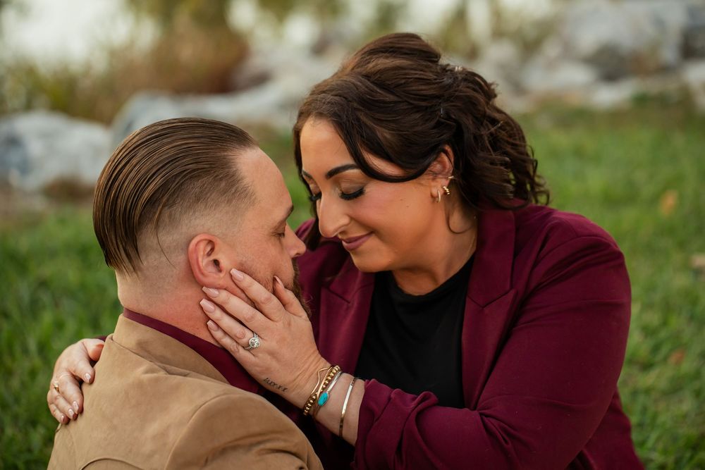 Couple sharing a romantic moment outdoors, woman in burgundy blazer tenderly touching mans face while smiling.