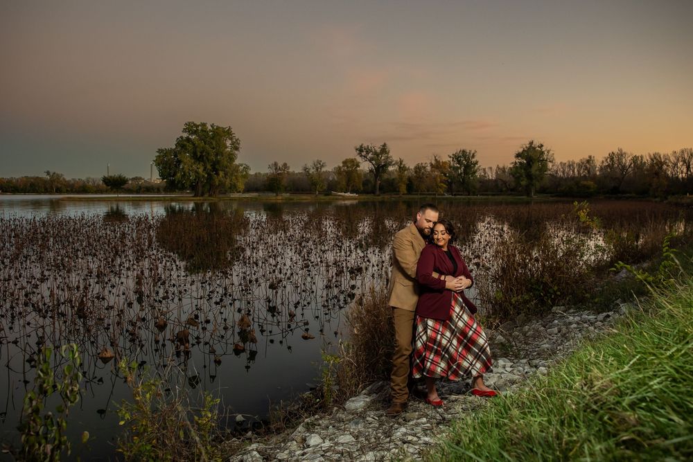 Couple embracing at sunset by a tranquil lake surrounded by autumn trees and tall grass in warm evening light.