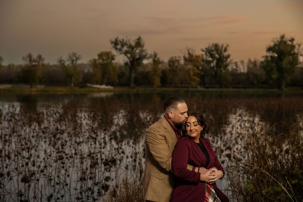 Couple embraces romantically at sunset near a serene wetland, woman in burgundy dress, man in tan blazer, autumn trees behind.