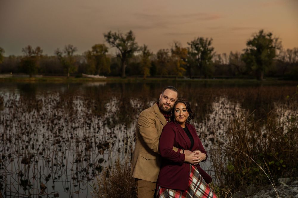 Couple embracing at sunset near a serene pond, surrounded by autumn trees, man in tan jacket, woman in burgundy coat.