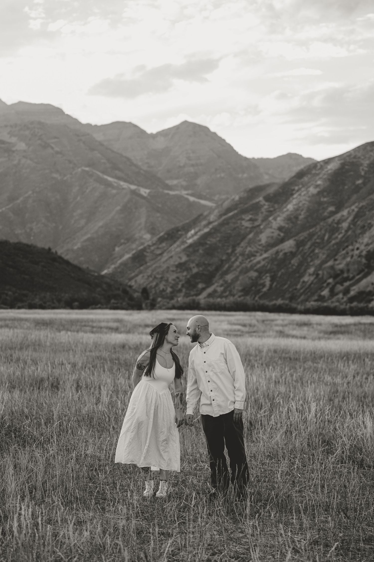 Black and white photo of a couple holding hands and sharing a kiss in a mountain meadow.