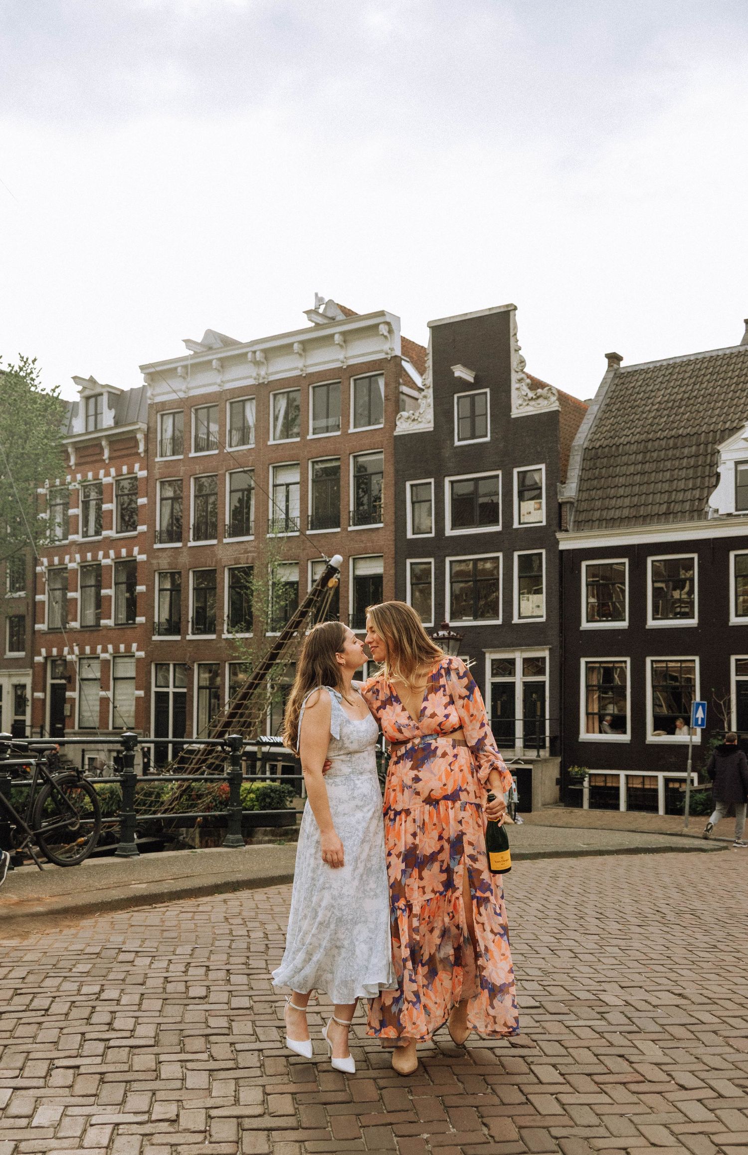 Two women in dresses share a kiss on a brick-paved canal bridge in Amsterdam, with historic gabled townhouses behind them.