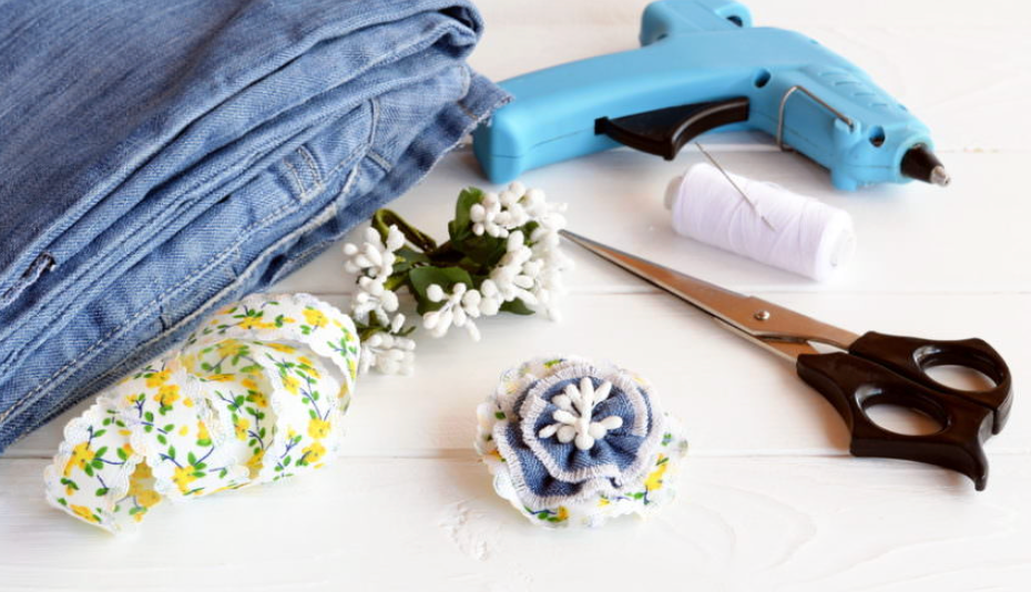 DIY craft supplies including denim fabric, glue gun, scissors, floral ribbon, and handmade fabric flower on white surface.