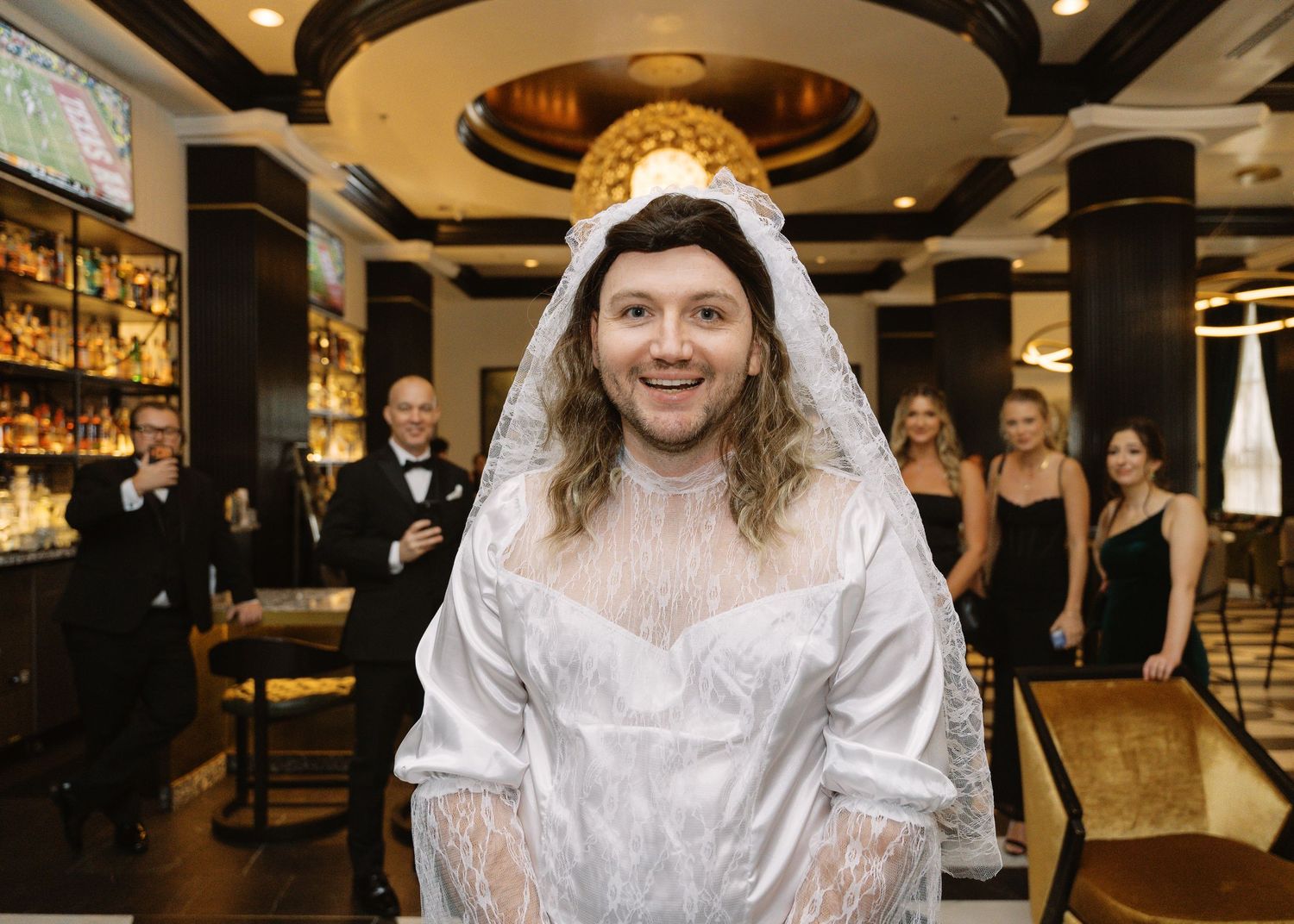 Person wearing a white wedding dress and veil smiling in an elegant bar setting with guests in formal attire behind.