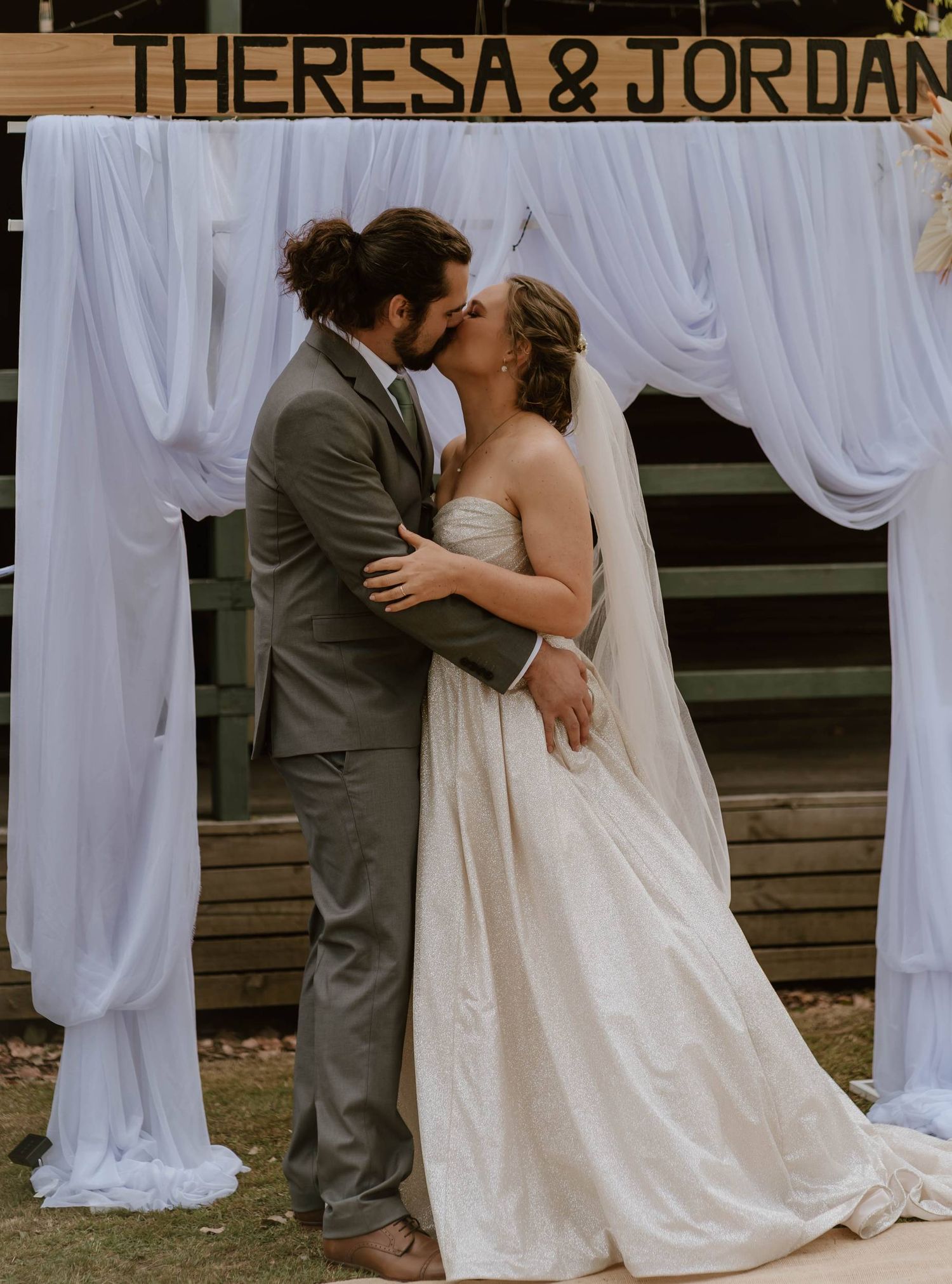 Theresa and Jordan share a first kiss as newlyweds under a white draped arch at their outdoor wedding ceremony.