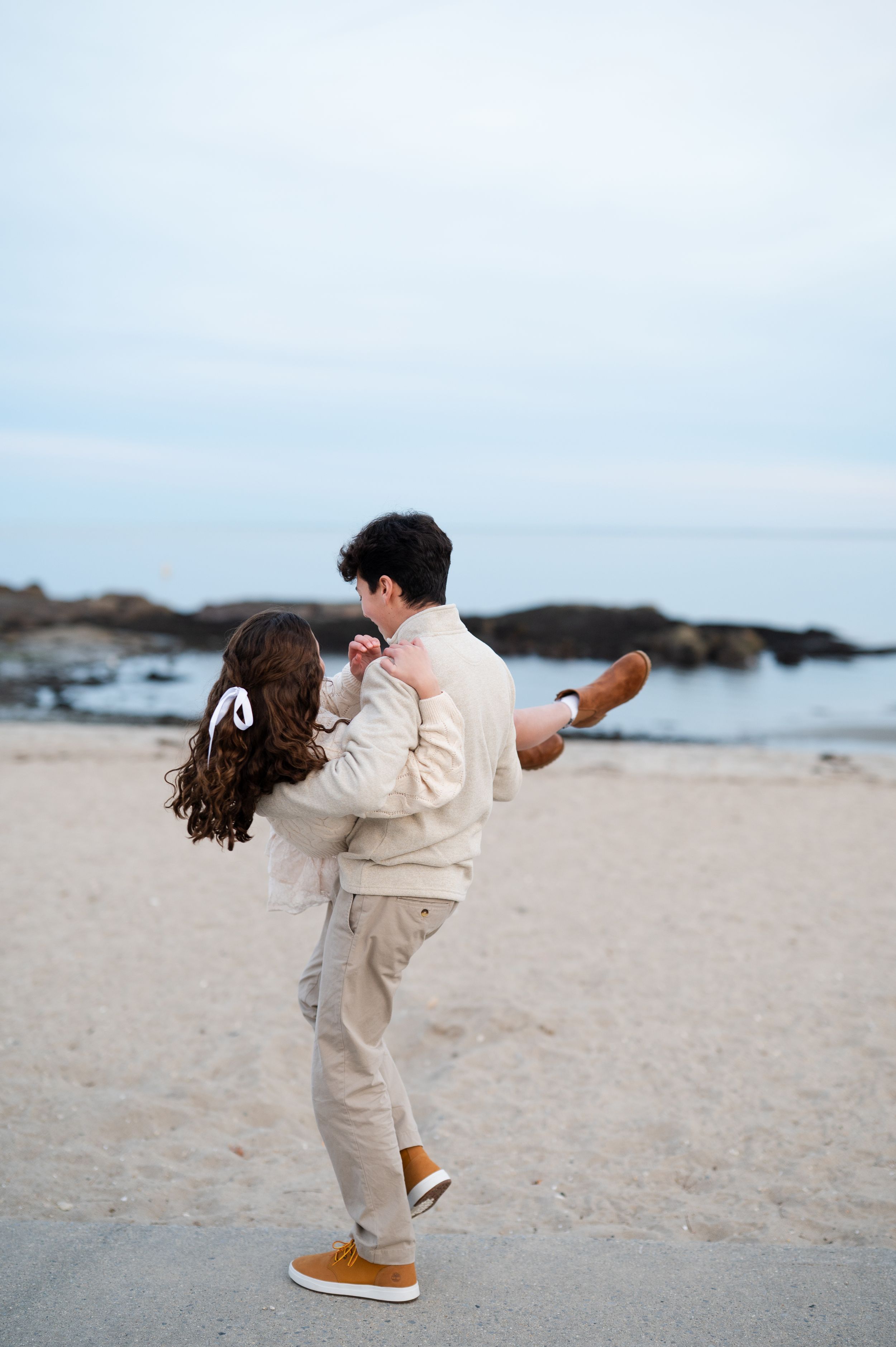 A brother and sister in cream sweaters pose together on a beach with calm water in the background.