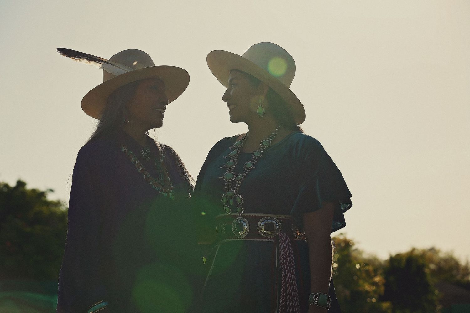 Two women in wide-brimmed hats, one with a feather, stand face to face in silhouette wearing dark garments and silver jewelry at sunset.