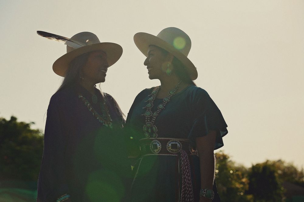 Two women in wide-brimmed hats, one with a feather, stand face to face in silhouette wearing dark garments and silver jewelry at sunset.
