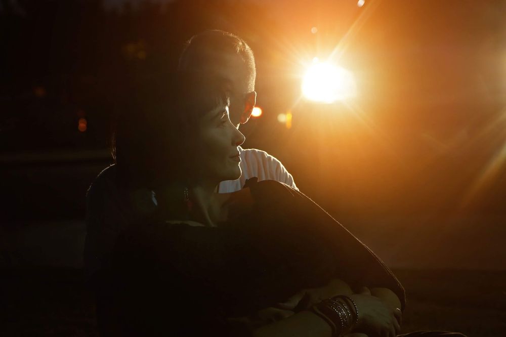 Silhouetted person in profile sits in dim light as a bright orange sun flare glows behind them, creating a moody, dramatic atmosphere.