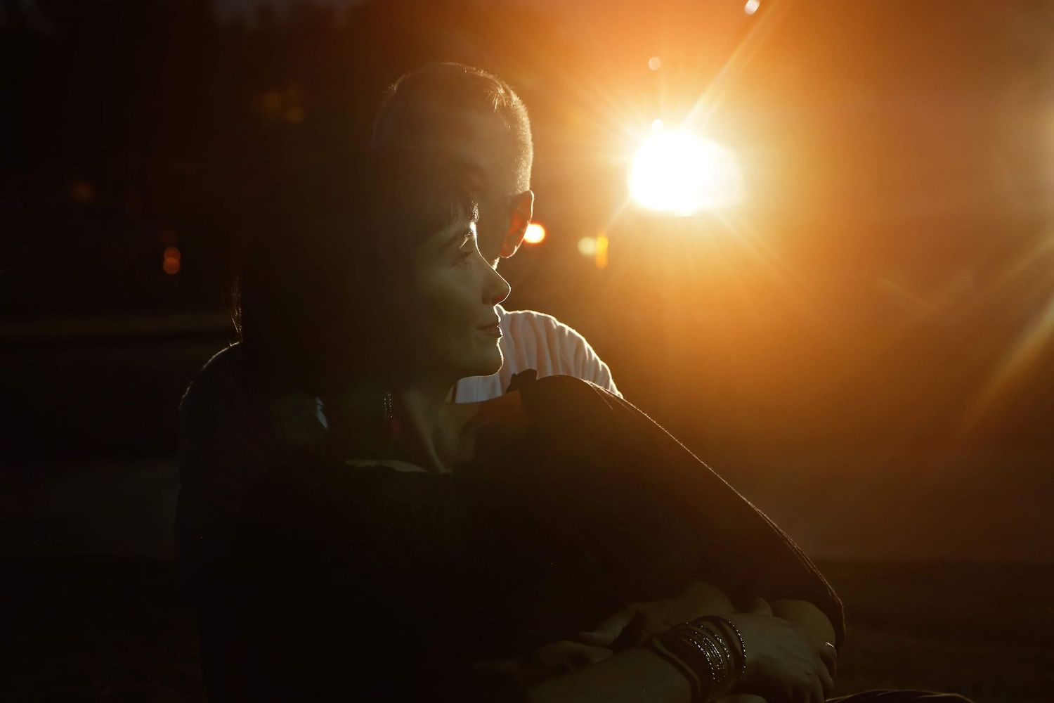 Silhouetted person in profile sits in dim light as a bright orange sun flare glows behind them, creating a moody, dramatic atmosphere.