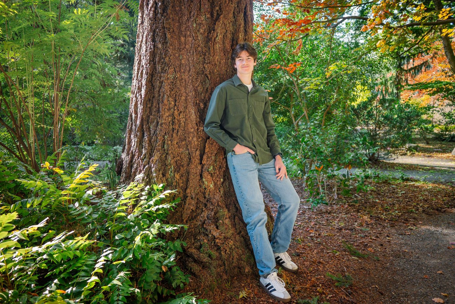 Senior leaning against a large tree surrounded by ferns and fall foliage