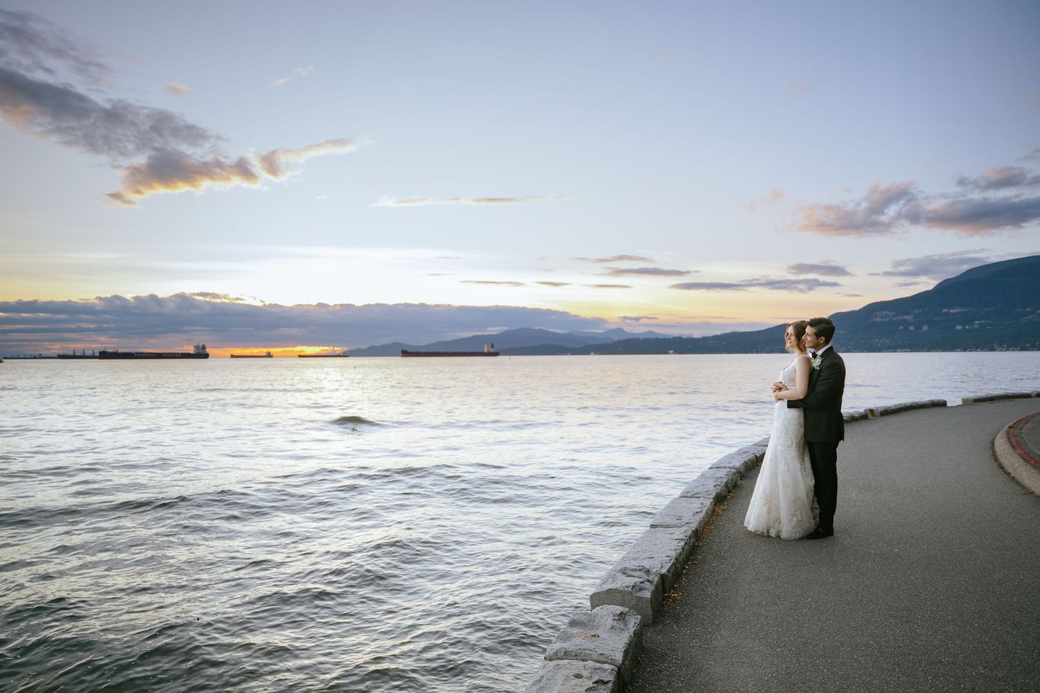Bride and groom stand by waterfront at sunset with mountains and ships in background.