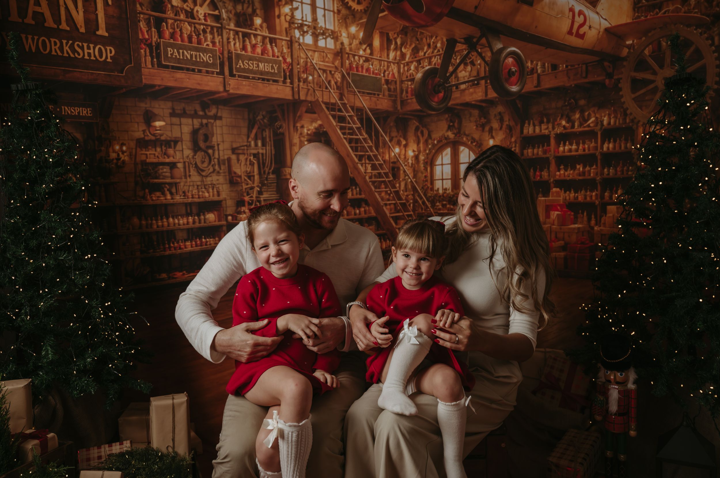 Family of four in cozy neutral and red outfits poses together in a festive Christmas workshop set with trees and wooden shelves.