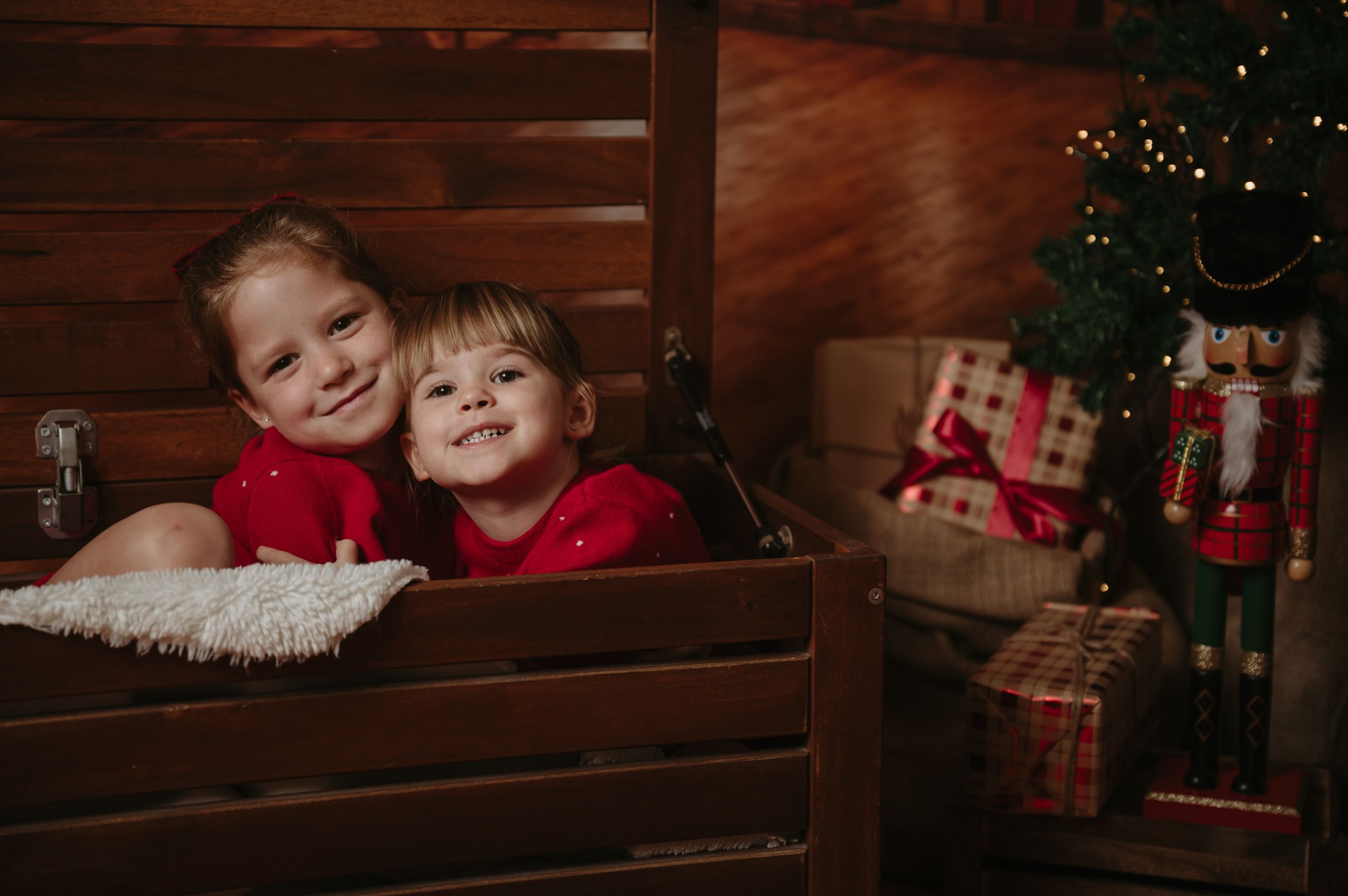 Two smiling children in red outfits pose together in a wooden crate beside a Christmas tree, nutcracker and wrapped gifts.