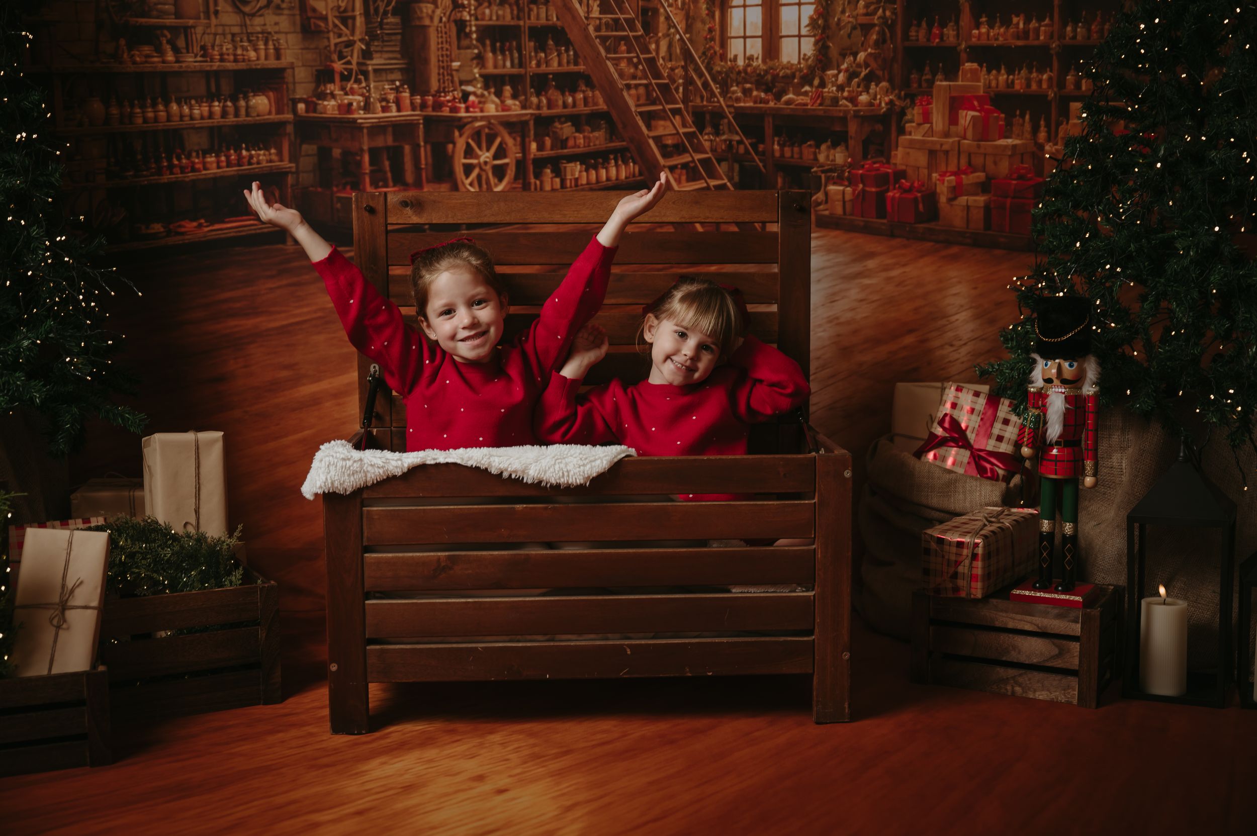 Two children in red outfits sit on a wooden bench with arms raised in a cozy Christmas studio set with gifts, a tree and candles.