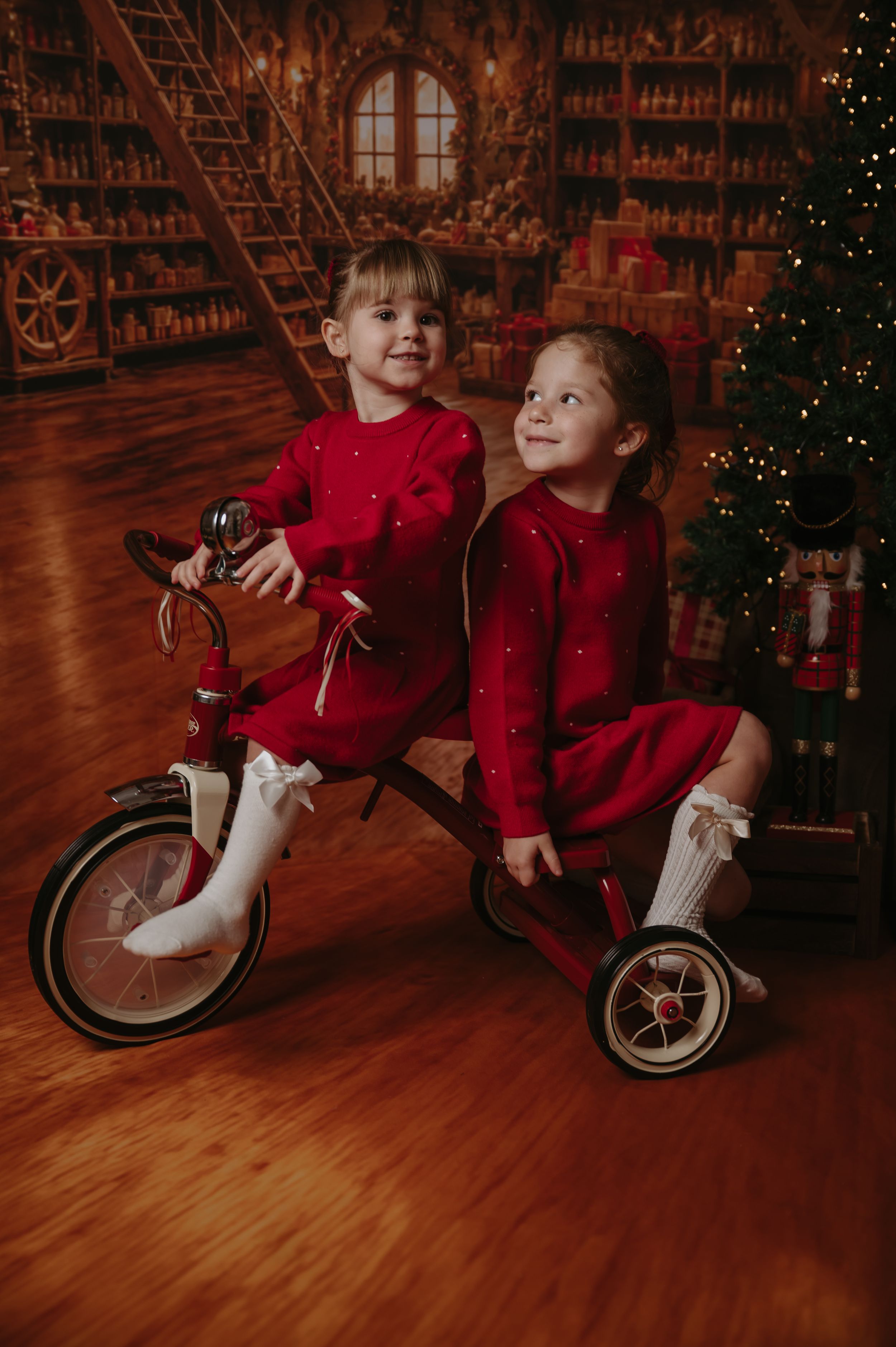 Two young girls in red dresses and white ribbon socks ride a vintage red tricycle in a cozy Christmas workshop with a decorated tree.