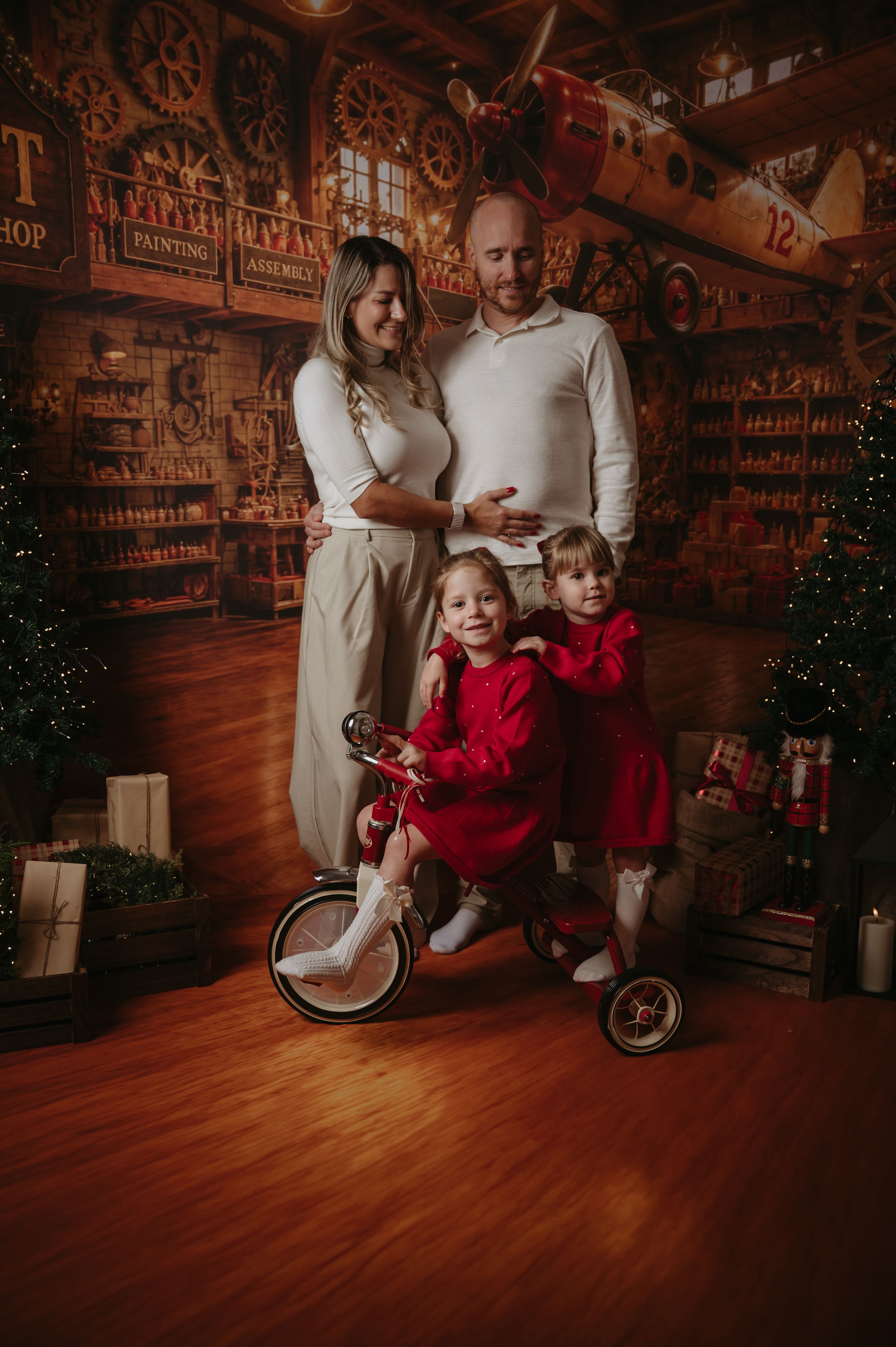 Family of four in a festive Christmas toy workshop studio, two girls in red dresses on a vintage red tricycle beside smiling parents.