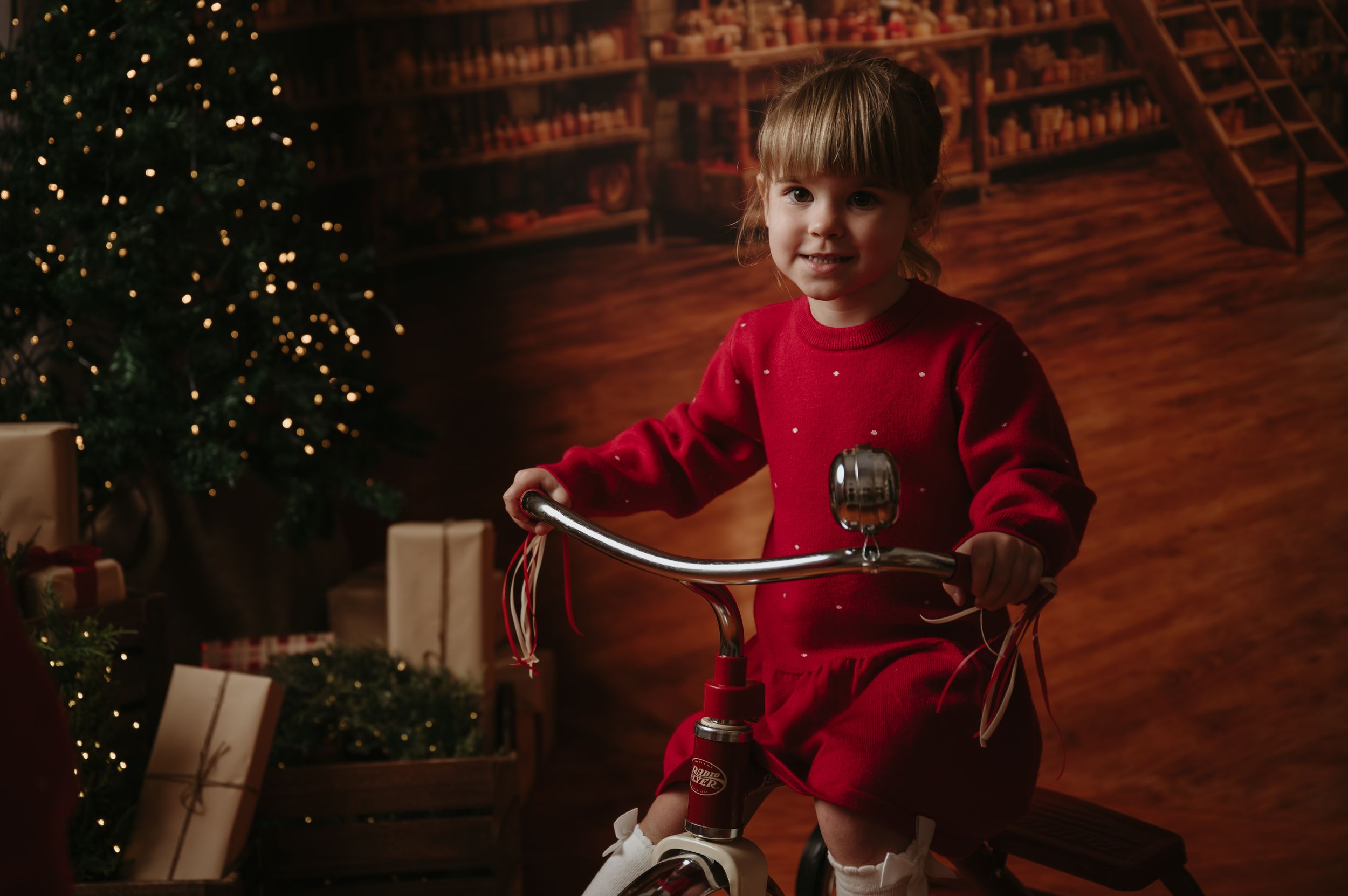 Smiling toddler in a red knit dress rides a vintage tricycle beside a lit Christmas tree and wrapped gifts in a cozy library setting.