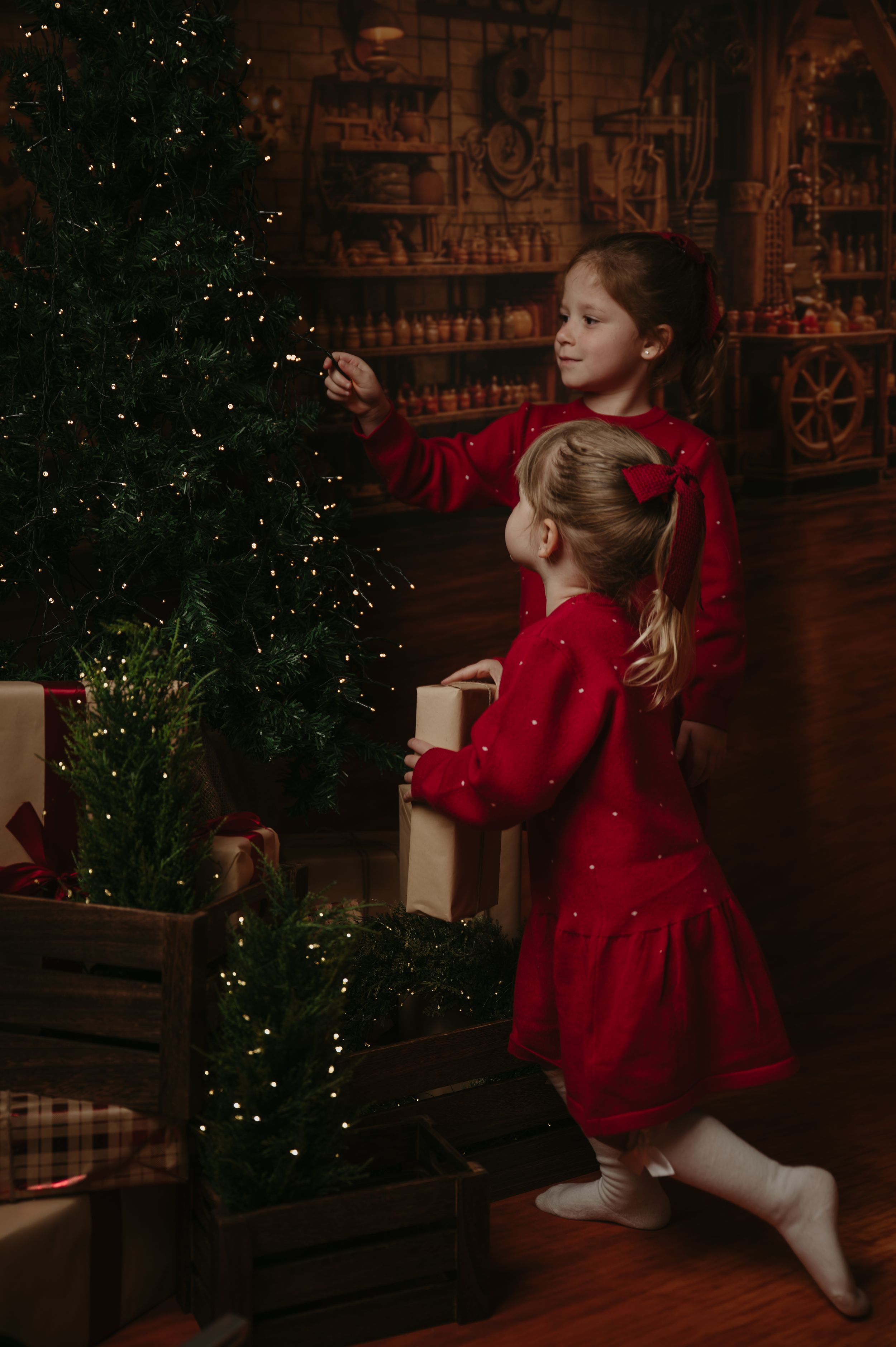 Two little girls in red dresses decorate a sparkling Christmas tree with twinkling lights in a cozy, rustic holiday workshop setting.