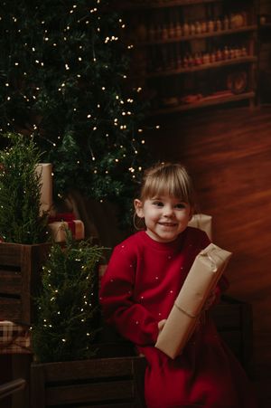 Smiling little girl in a red sweater dress holds a kraft-wrapped Christmas gift beside a lit tree in a cozy wooden holiday setting.