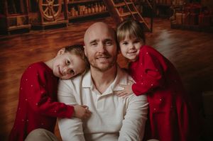 Smiling bald man in a white shirt sits with two young children in red outfits leaning on his shoulders in a cozy, rustic holiday setting.