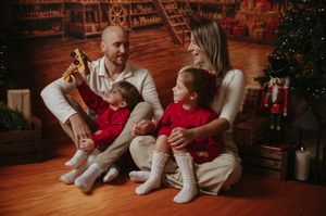Smiling parents in white sit on a wooden floor with two toddlers in red outfits, playing with a toy airplane beside a Christmas tree.