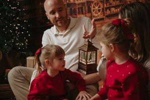 Man and two young girls in matching red outfits gather around a glowing lantern beside a Christmas tree in a cozy holiday setting.