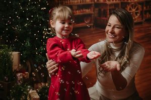 Smiling woman and young girl in a red dress play with falling bubbles beside a decorated Christmas tree in a cozy, warmly lit room.