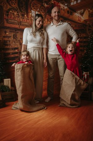 Smiling parents stand with two young children in burlap sacks in a cozy vintage Christmas toy workshop for a festive family photo.