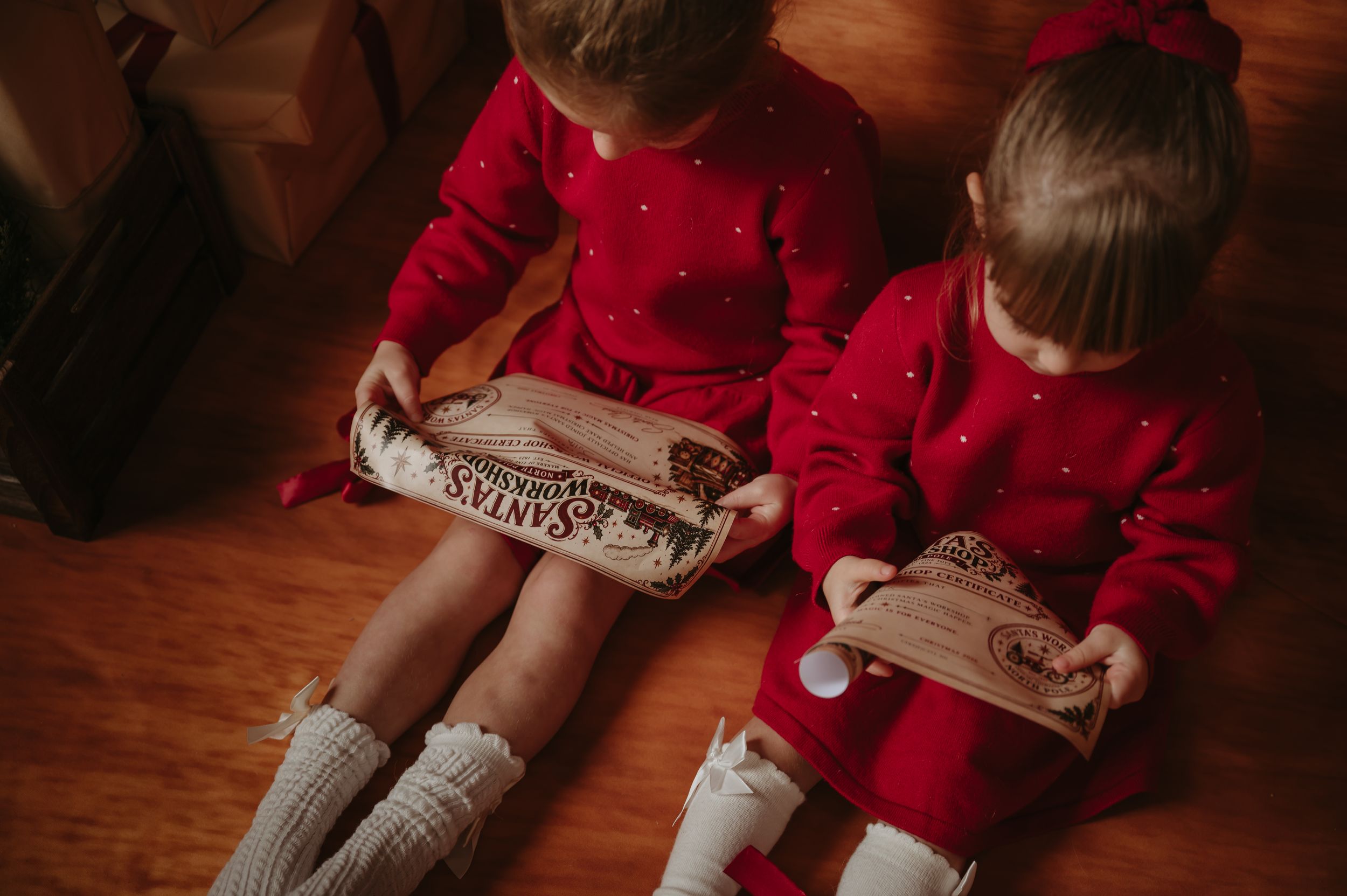 Two little girls in matching red dotted dresses and white knit socks sit on a wooden floor opening Christmas gifts.