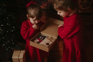 Two young girls in red dresses look into an open kraft gift box holding a photo and keepsakes beside a lit Christmas tree.