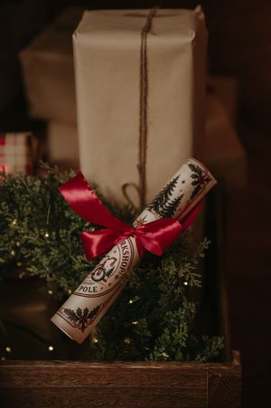 Festive Christmas cracker tied with a red satin bow rests on evergreen branches with twinkling lights beside a kraft paper wrapped gift.